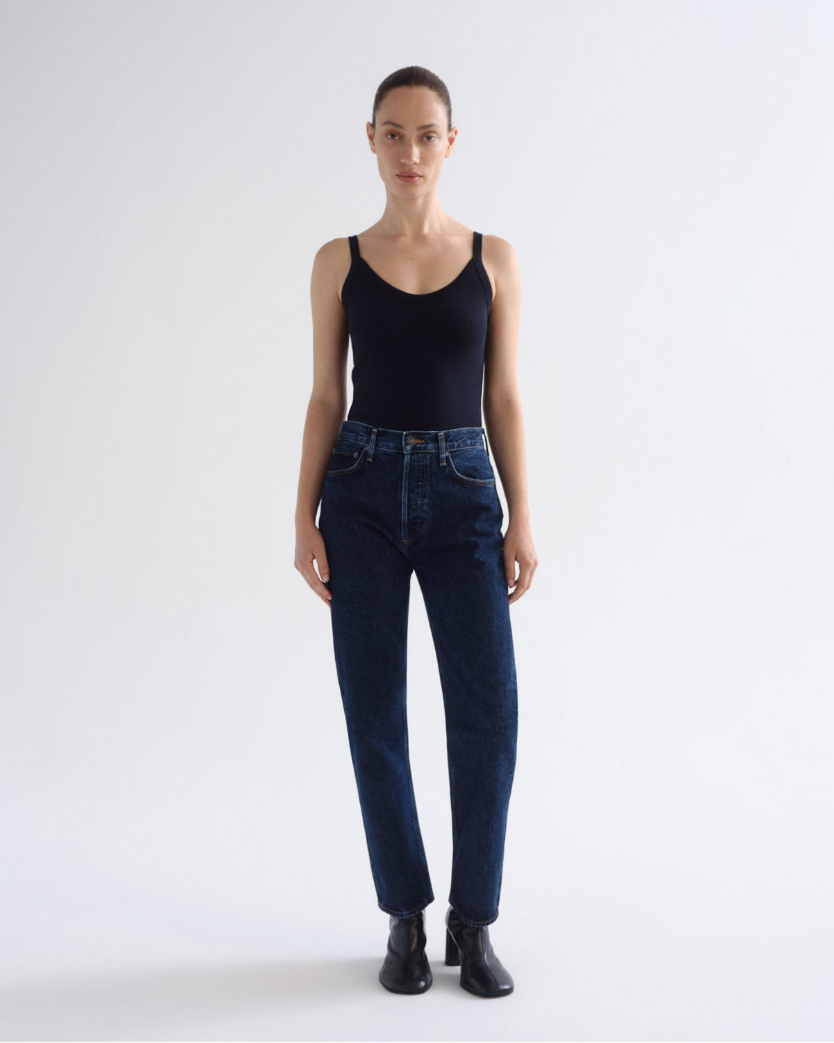 A model stands against a light gray studio background wearing a black ribbed tank top, dark blue high-waisted straight-leg jeans, and black ankle boots.