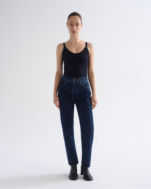 A model stands against a light gray studio background wearing a black ribbed tank top, dark blue high-waisted straight-leg jeans, and black ankle boots.