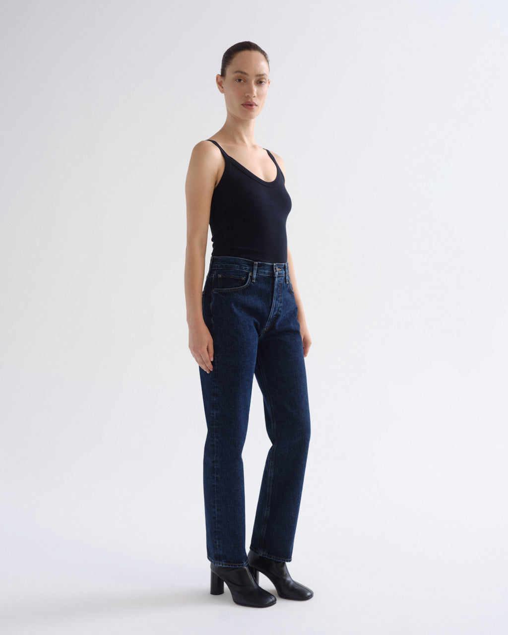 A model stands in profile wearing dark blue straight-leg jeans, a black tank top, and black ankle boots against a plain light gray studio background.