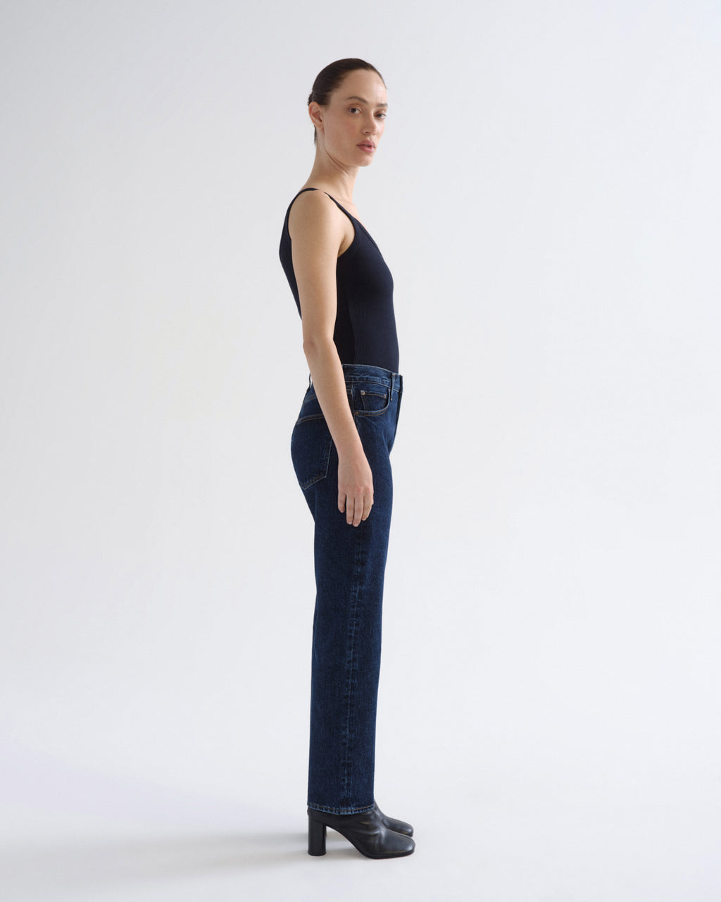 A model is shown in profile wearing dark blue straight-leg jeans, a black sleeveless top, and black high-heeled shoes against a plain white studio background.