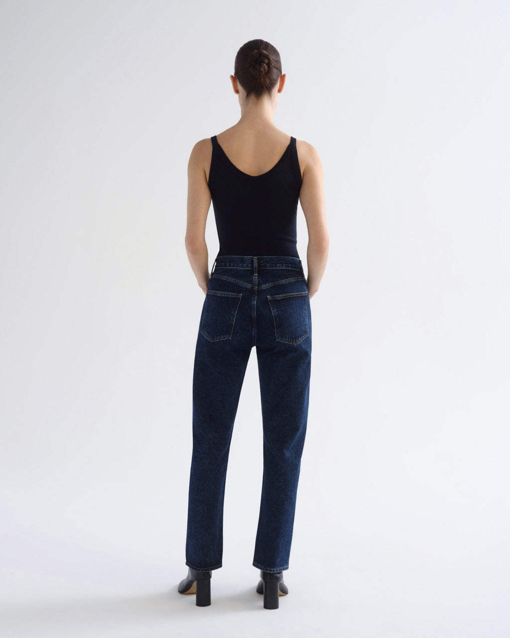 A model viewed from behind wearing a black tank top and dark blue straight-leg jeans, standing against a plain white studio background.