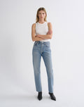 A model stands against a light gray studio background wearing a white ribbed sleeveless top, light-wash blue high-rise jeans, and black pointed ankle boots.