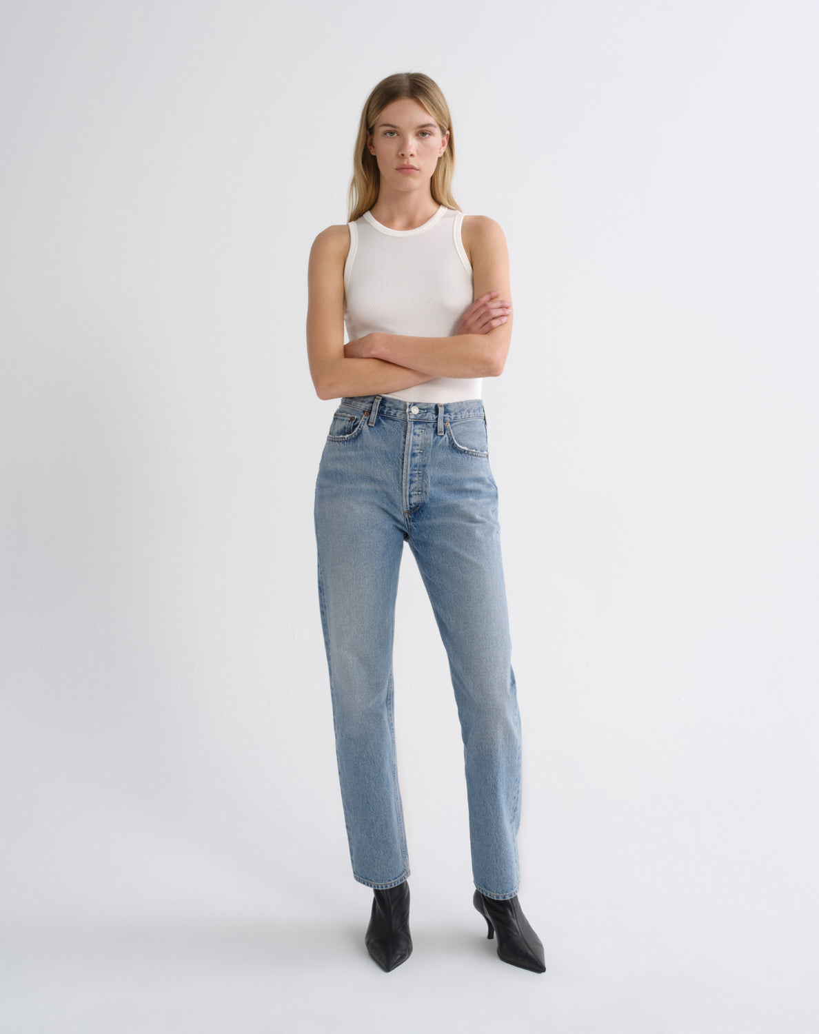 A model stands against a light gray studio background wearing a white ribbed sleeveless top, light-wash blue high-rise jeans, and black pointed ankle boots.