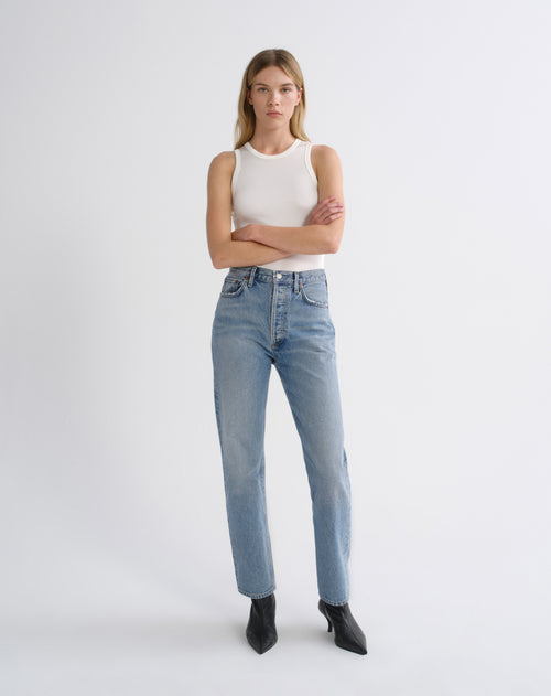 A model stands against a light gray studio background wearing a white ribbed sleeveless top, light-wash blue high-rise jeans, and black pointed ankle boots.