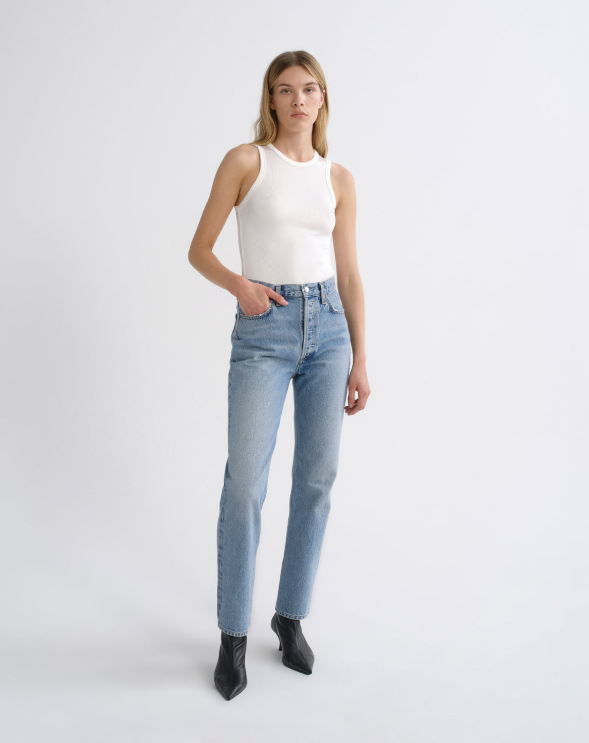 A woman modeling a white ribbed sleeveless top and light-wash blue straight-leg jeans with black ankle boots, standing against a light gray studio background.