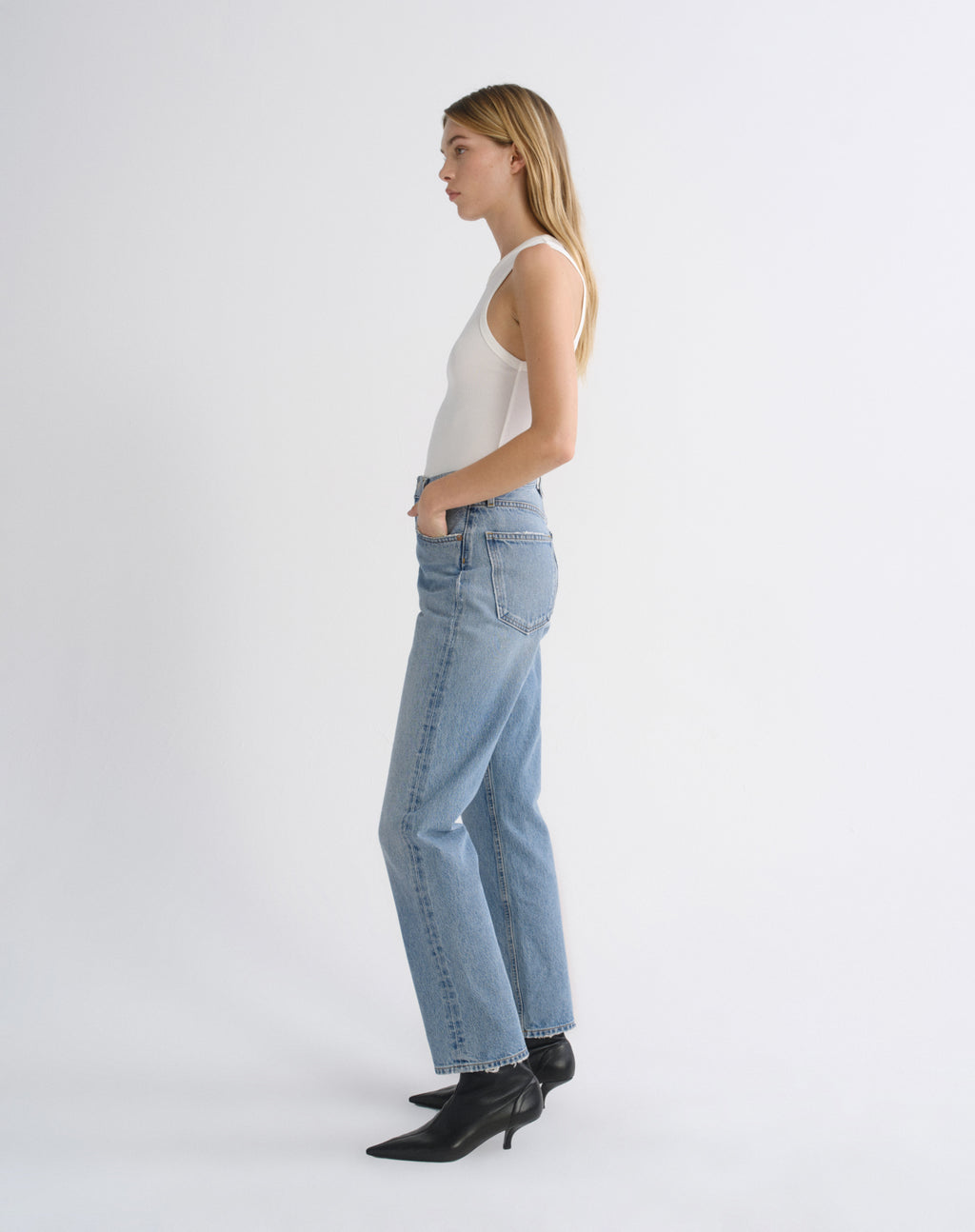 A woman in a white tank top, light-wash blue jeans, and black pointed-toe ankle boots is shown in profile against a clean white studio background.