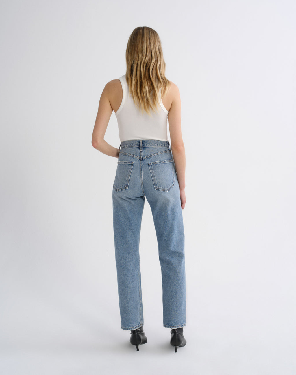 A woman is shown from behind wearing light-wash blue straight-leg jeans, a white ribbed tank top, and black pointed-toe heels against a plain light gray studio background.