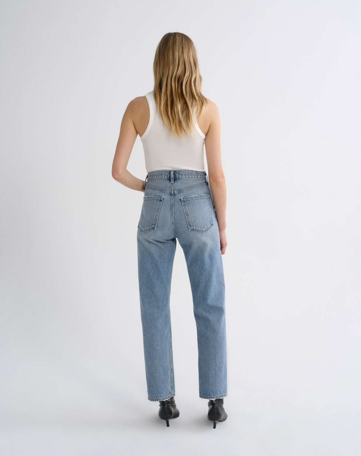 A woman is shown from behind wearing light-wash blue straight-leg jeans, a white ribbed tank top, and black pointed-toe heels against a plain light gray studio background.