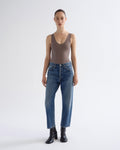 A model stands against a light gray studio background wearing a fitted taupe-brown sleeveless bodysuit, blue straight-leg jeans, and black ankle boots.