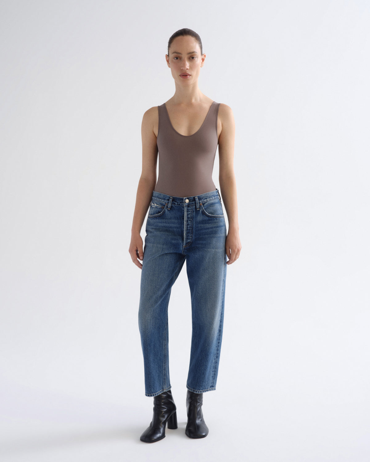 A model stands against a light gray studio background wearing a fitted taupe-brown sleeveless bodysuit, blue straight-leg jeans, and black ankle boots.