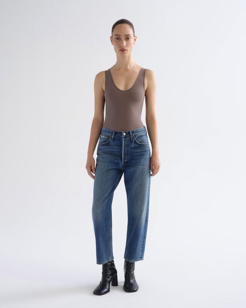 A model stands against a light gray studio background wearing a fitted taupe-brown sleeveless bodysuit, blue straight-leg jeans, and black ankle boots.