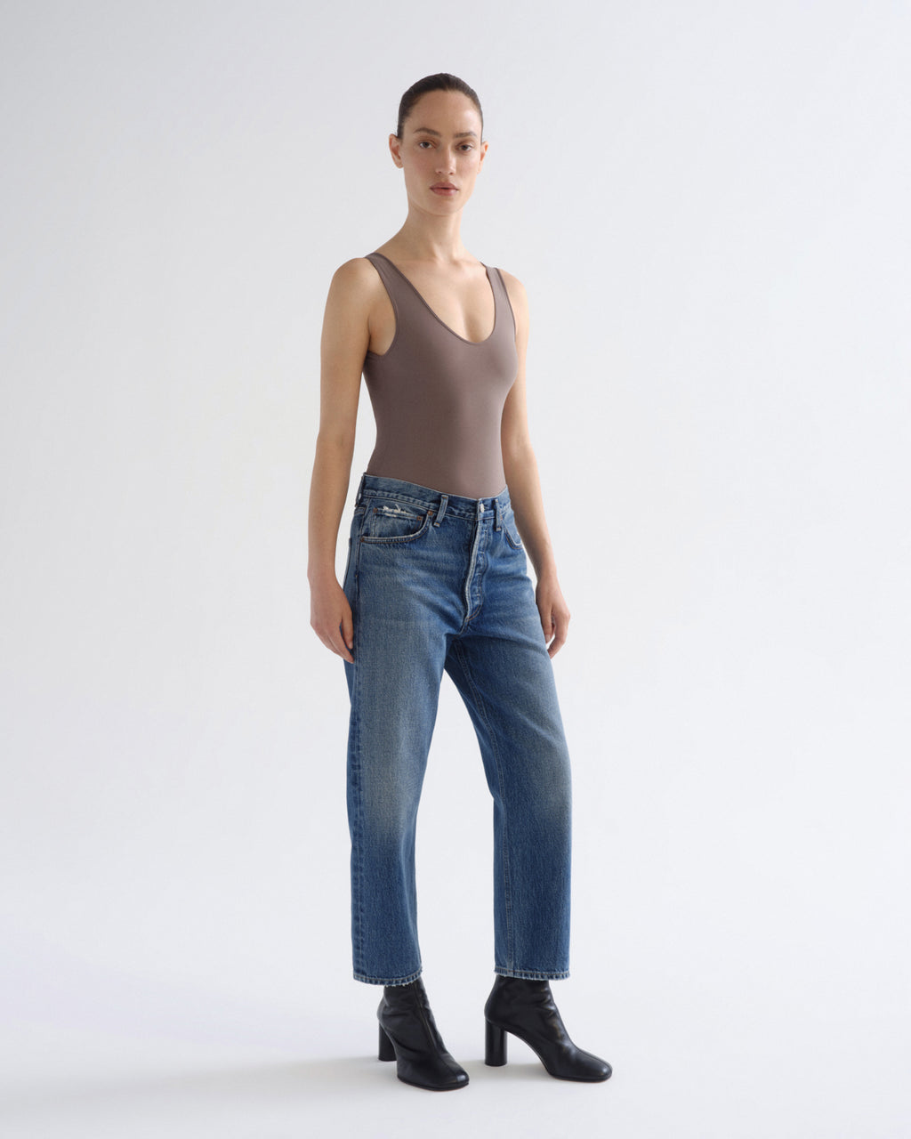 A model stands against a light gray studio background wearing a taupe-brown ribbed tank top, blue jeans, and black ankle boots.