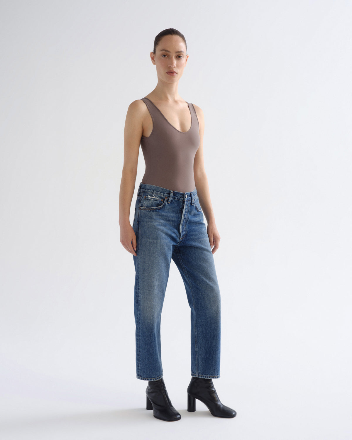 A model stands against a light gray studio background wearing a taupe-brown ribbed tank top, blue jeans, and black ankle boots.