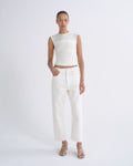 A model stands against a light gray studio background wearing a sleeveless white ribbed crop top, white cropped jeans, and clear-strap heeled sandals.