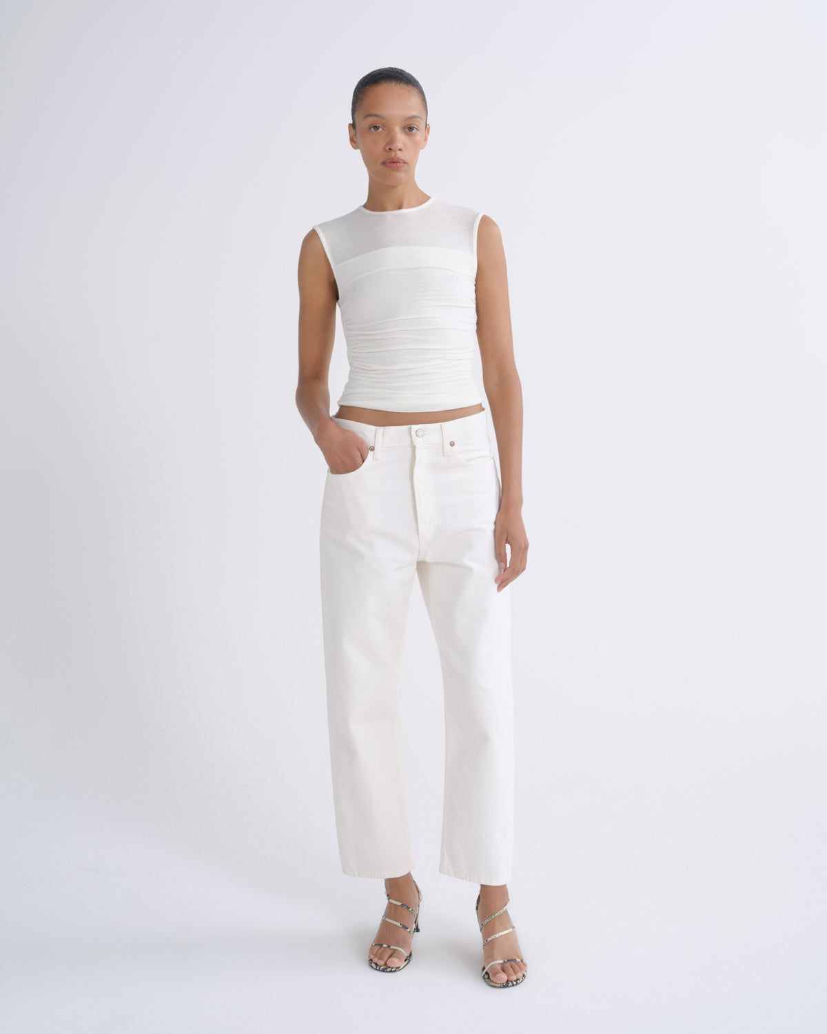 A model stands against a light gray studio background wearing a sleeveless white ribbed crop top, white cropped jeans, and clear-strap heeled sandals.