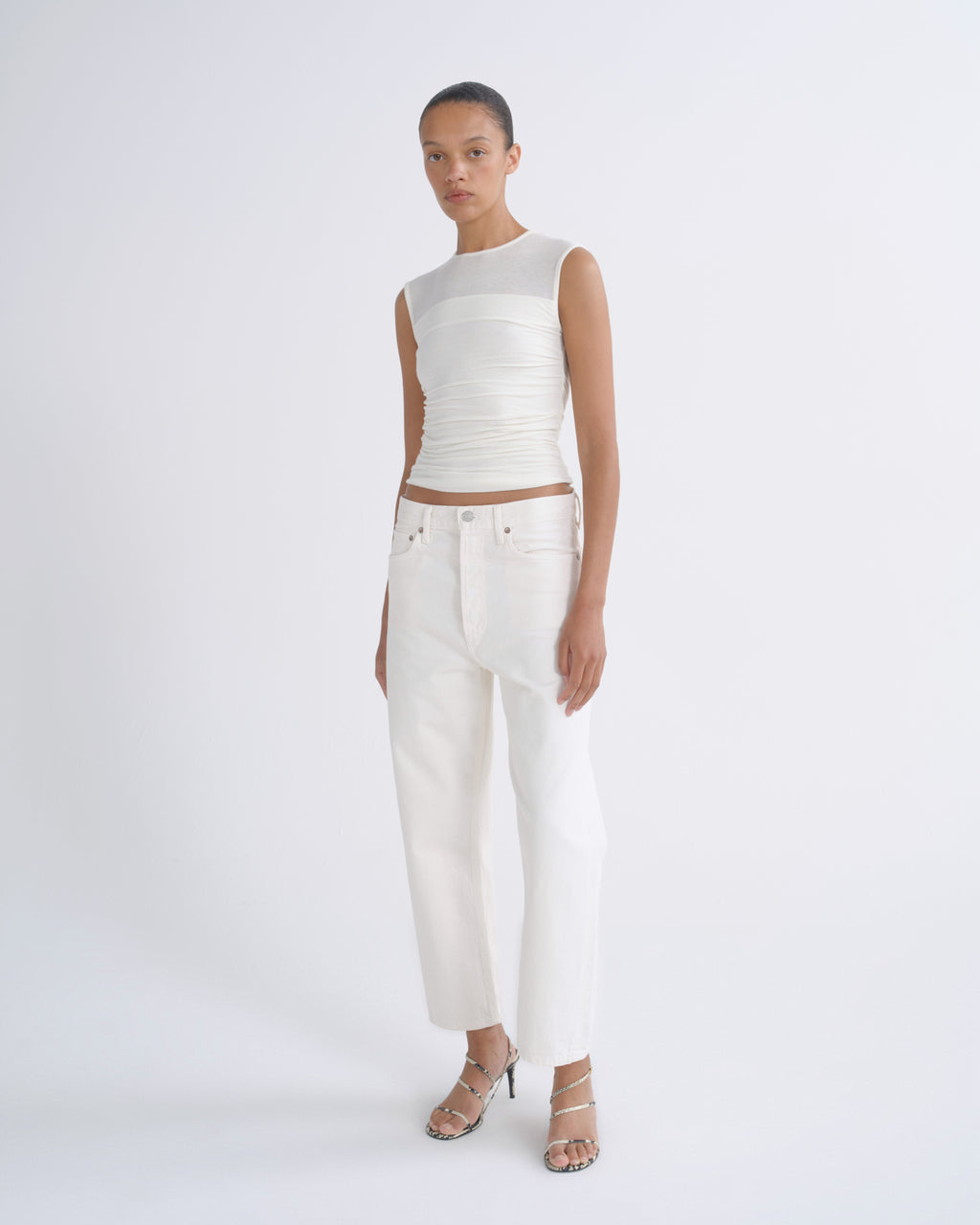 A model stands against a light gray studio background wearing a white sleeveless top with a sheer mesh yoke and gathered waist, paired with white cropped pants and clear-strap high heels.