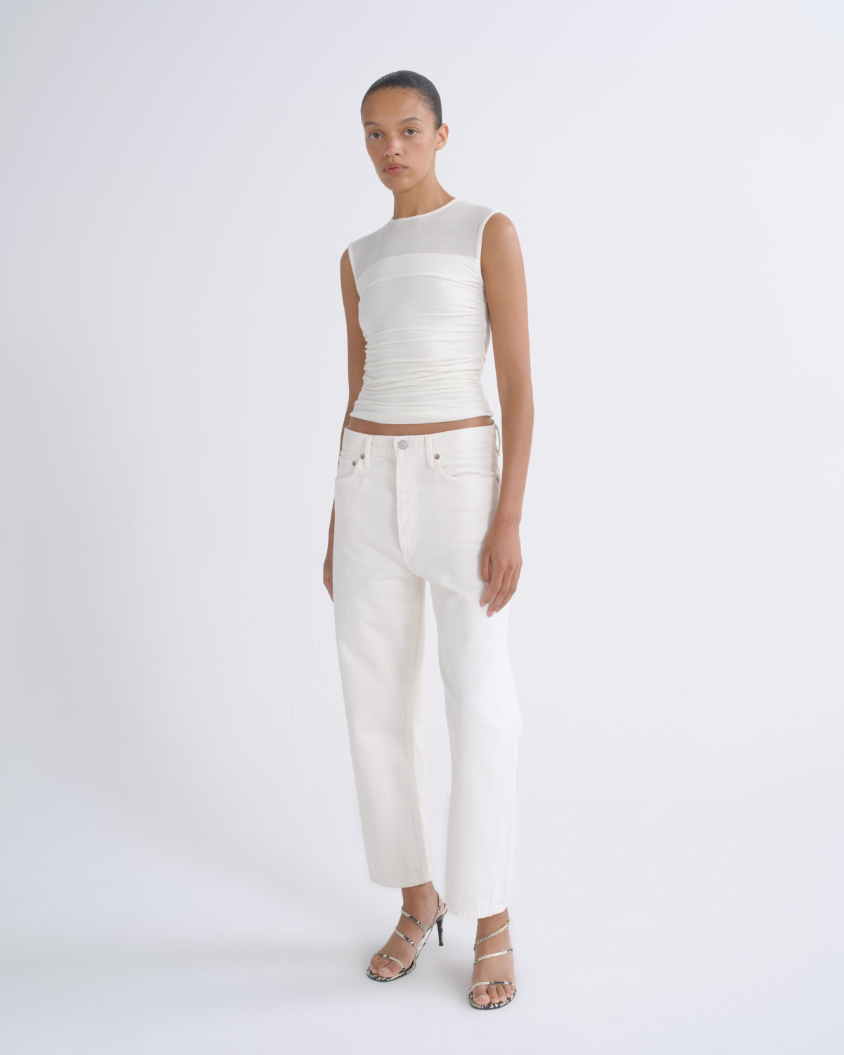 A model stands against a light gray studio background wearing a white sleeveless top with a sheer mesh yoke and gathered waist, paired with white cropped pants and clear-strap high heels.