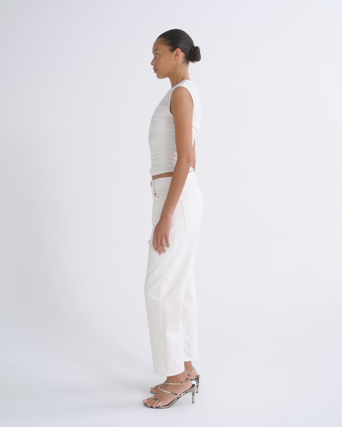 A woman stands in profile wearing a white ribbed sleeveless crop top, white wide-leg pants, and metallic heeled sandals against a clean white studio background.