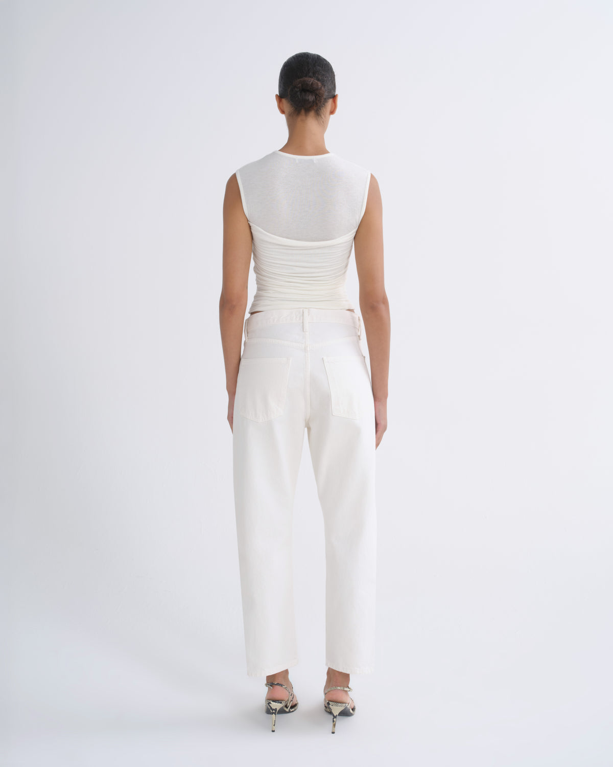 A model viewed from the back wearing a white sleeveless textured top and white wide-leg trousers, paired with metallic high-heeled sandals, against a plain white studio background.