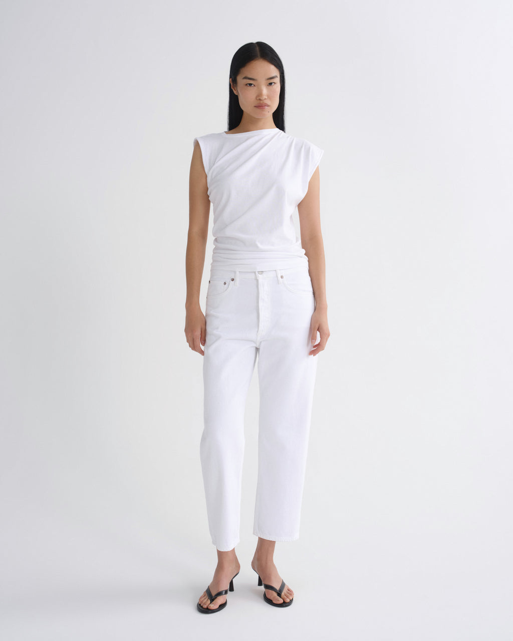 A model stands against a light gray studio background wearing a white asymmetrical sleeveless top, white cropped pants, and black sandals.