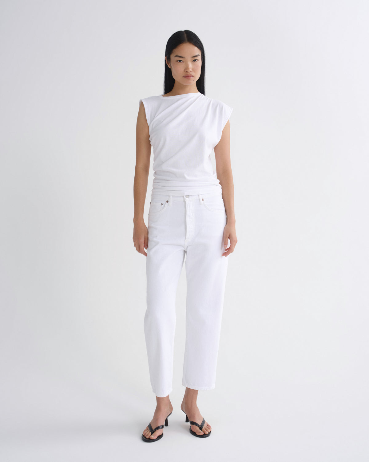 A model stands against a light gray studio background wearing a white asymmetrical sleeveless top, white cropped pants, and black sandals.