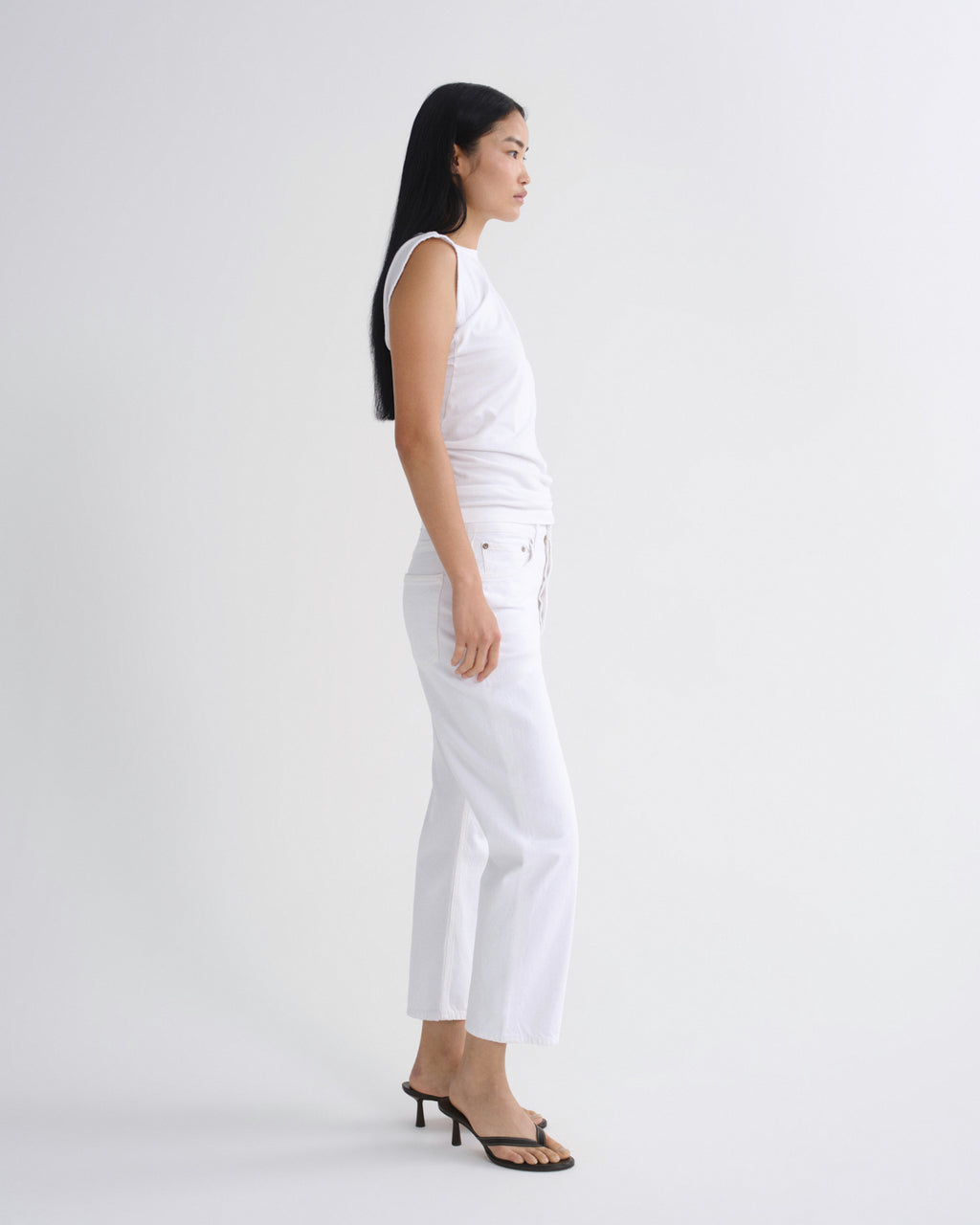 A woman stands in profile wearing a sleeveless white top and white cropped pants with black heeled sandals against a plain light gray studio background.
