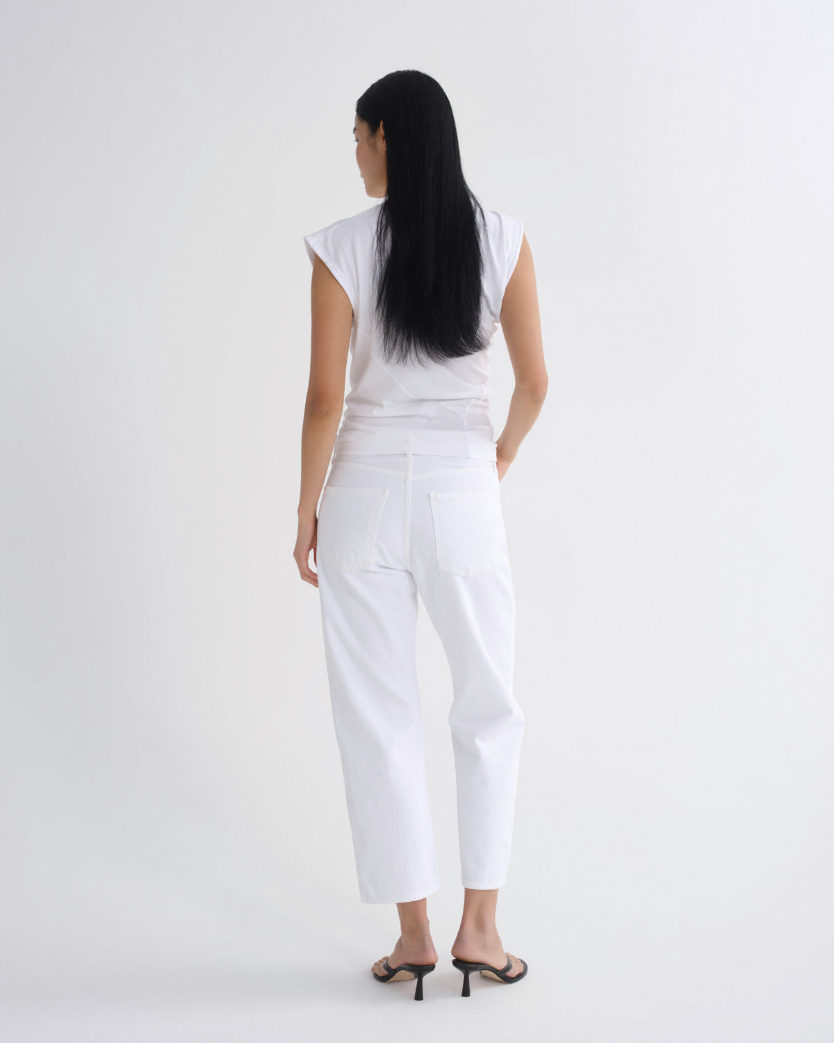 A woman viewed from the back wearing a white sleeveless top and white cropped pants, standing against a plain light gray studio background.