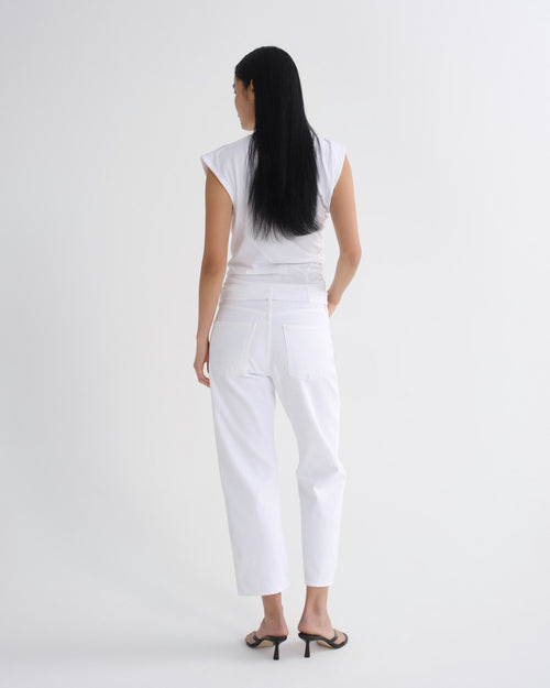 A woman viewed from the back wearing a white sleeveless top and white cropped pants, standing against a plain light gray studio background.