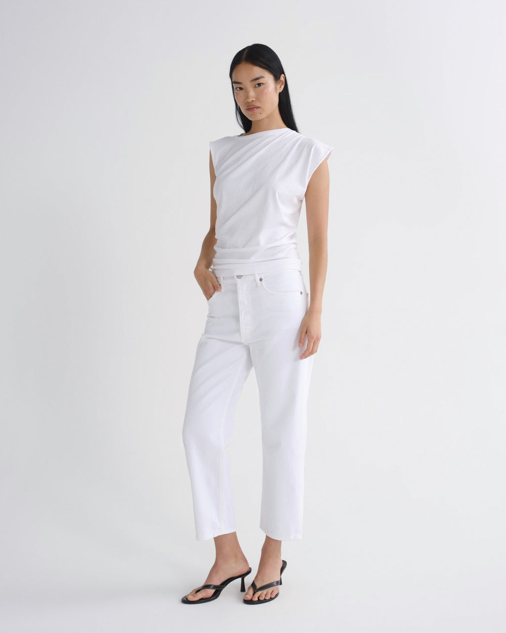 A model wearing a white draped sleeveless top and white cropped jeans with black sandals, standing against a plain light gray studio background.