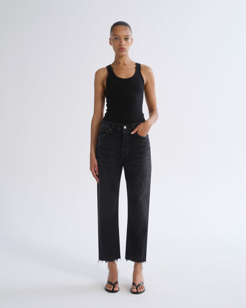 A model stands against a light gray studio background wearing a black ribbed tank top, black cropped wide-leg jeans with raw hems, and black sandals.