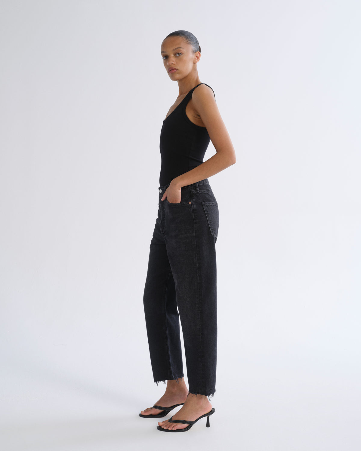 A model wearing a black sleeveless top, black wide-leg cropped jeans, and black strappy high-heeled sandals, standing in profile against a plain light gray studio background.