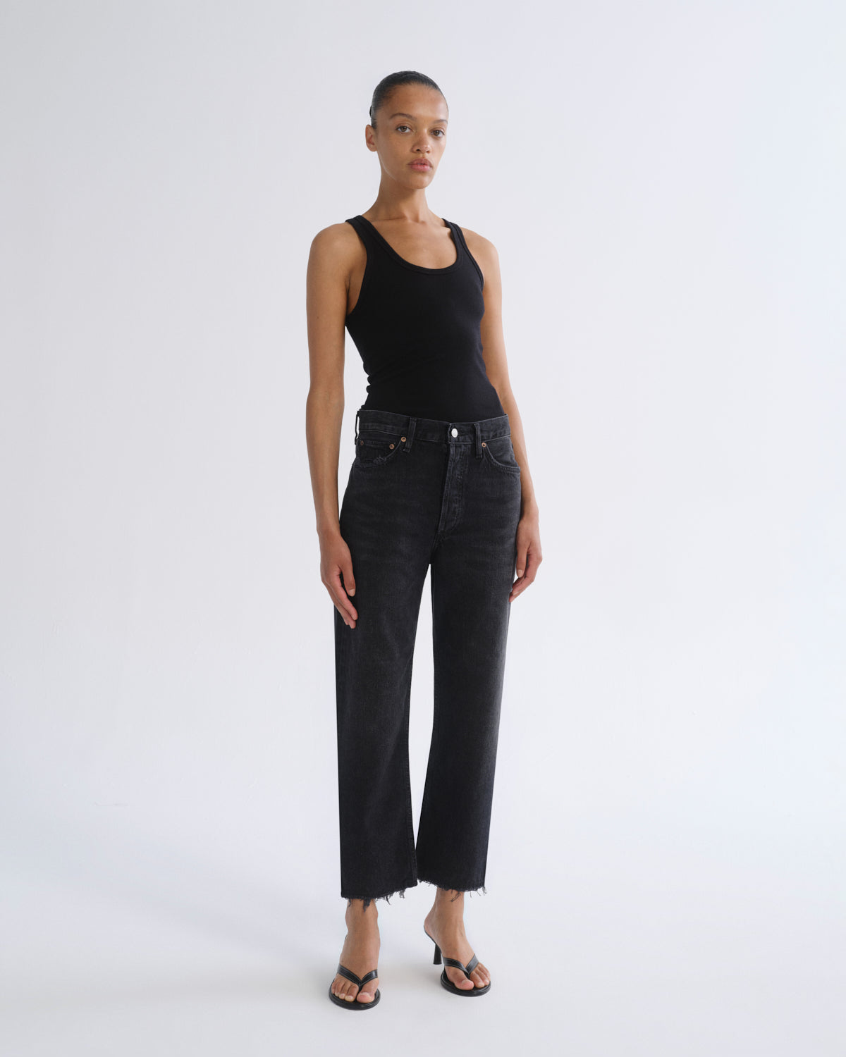 A model stands against a light gray studio background wearing a black ribbed tank top, black wide-leg cropped jeans with a raw hem, and black sandals.