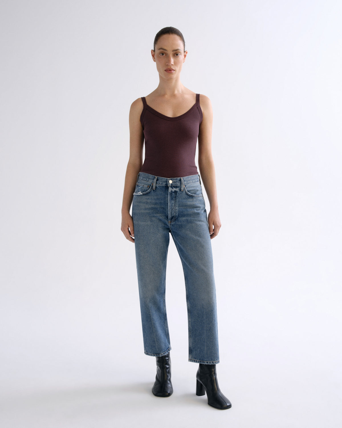 A model stands against a light gray studio background wearing a dark burgundy camisole, blue straight-leg jeans, and black ankle boots.