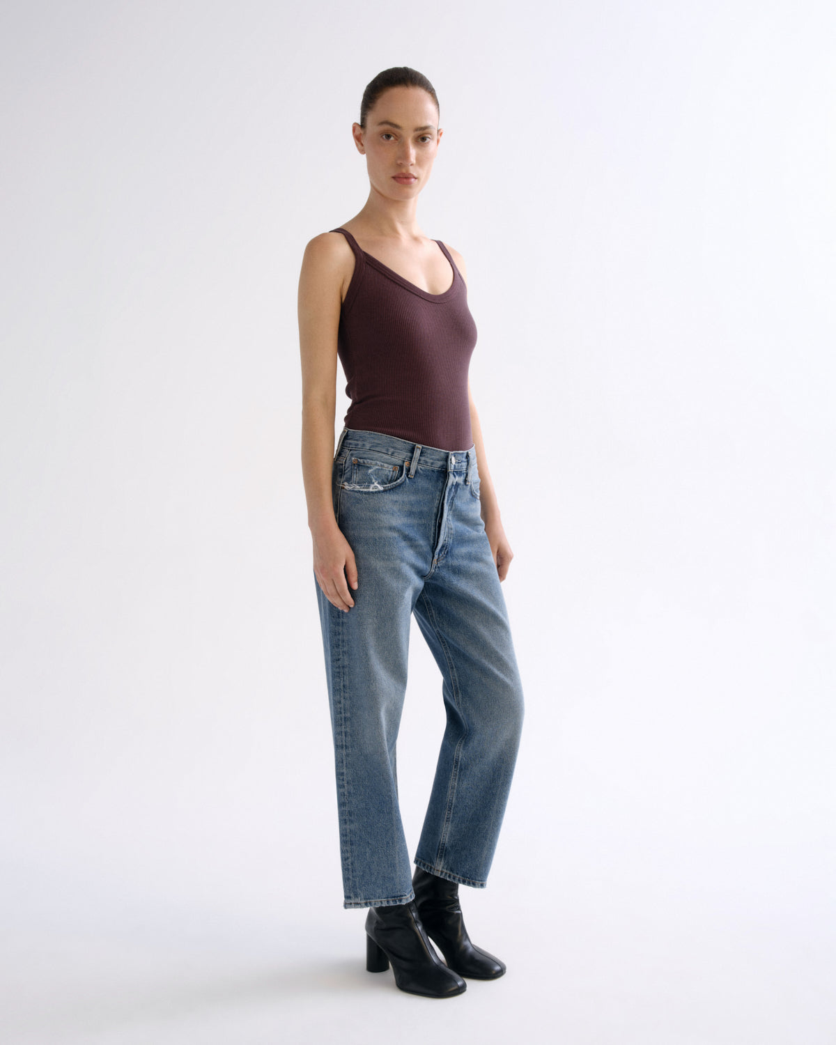 A model stands against a plain white studio background wearing a dark plum sleeveless tank top, light-wash blue straight-leg jeans, and black ankle boots.
