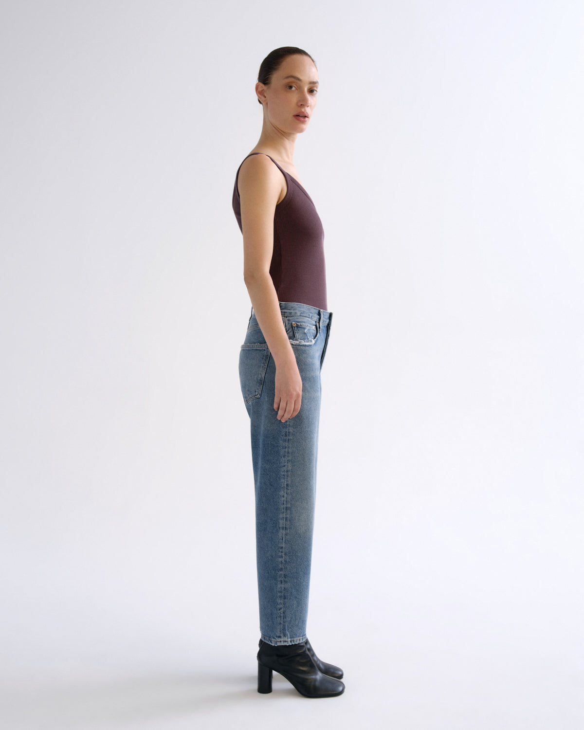 A woman stands in profile wearing a dark plum sleeveless top, light blue straight-leg jeans, and black ankle boots against a plain light gray studio background.