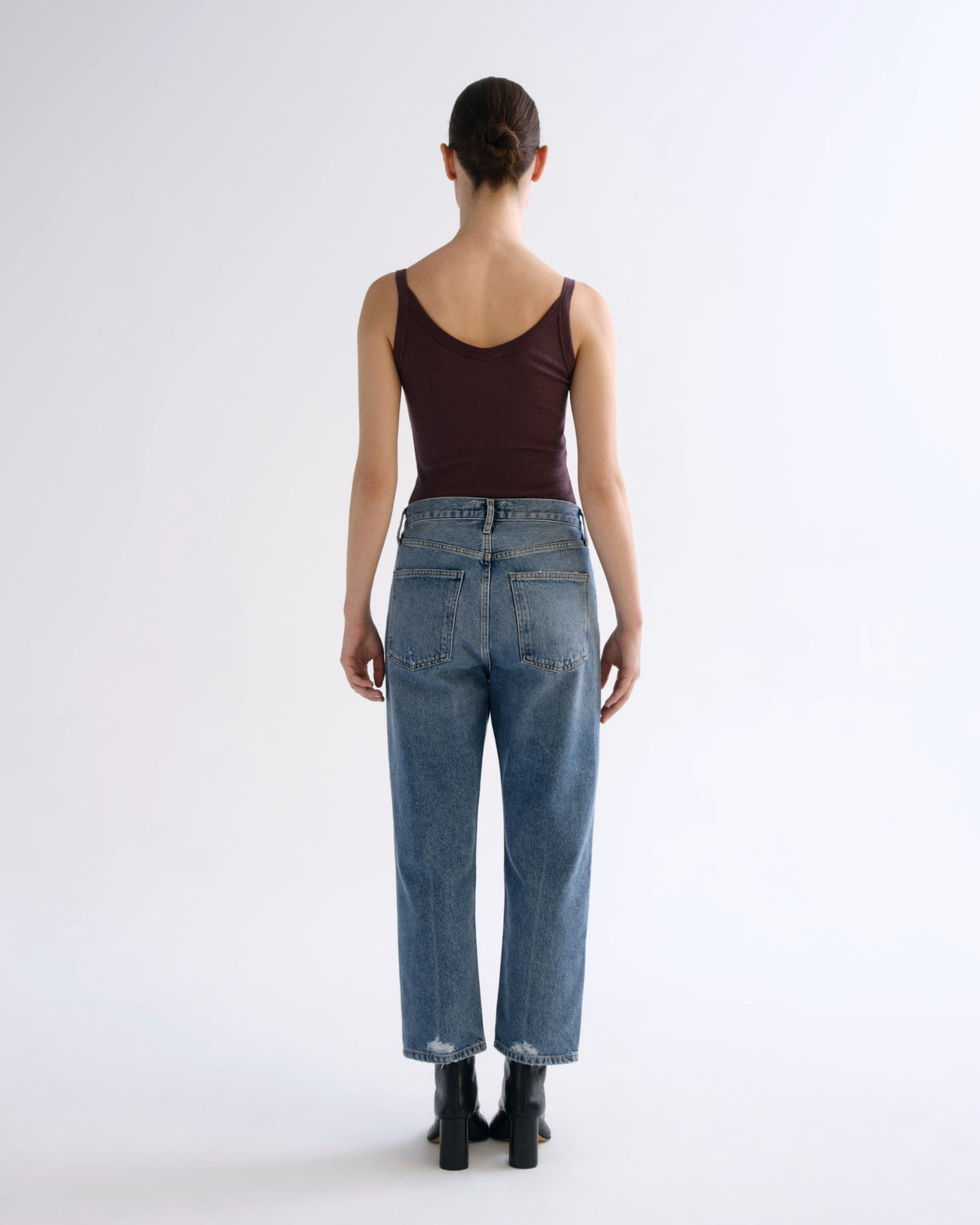 A model stands against a plain white studio background wearing a dark plum sleeveless tank top, light-wash blue straight-leg jeans, and black ankle boots.
