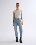 A model stands against a light gray studio background wearing a beige short-sleeve crewneck top, light-wash blue jeans, and black ankle boots.