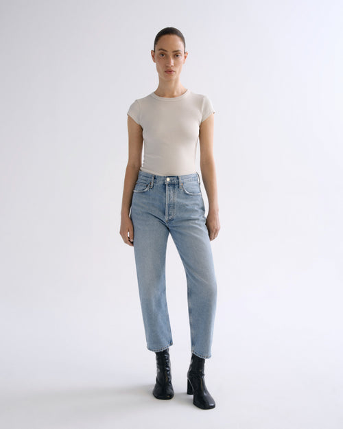 A model stands against a light gray studio background wearing a beige short-sleeve crewneck top, light-wash blue jeans, and black ankle boots.
