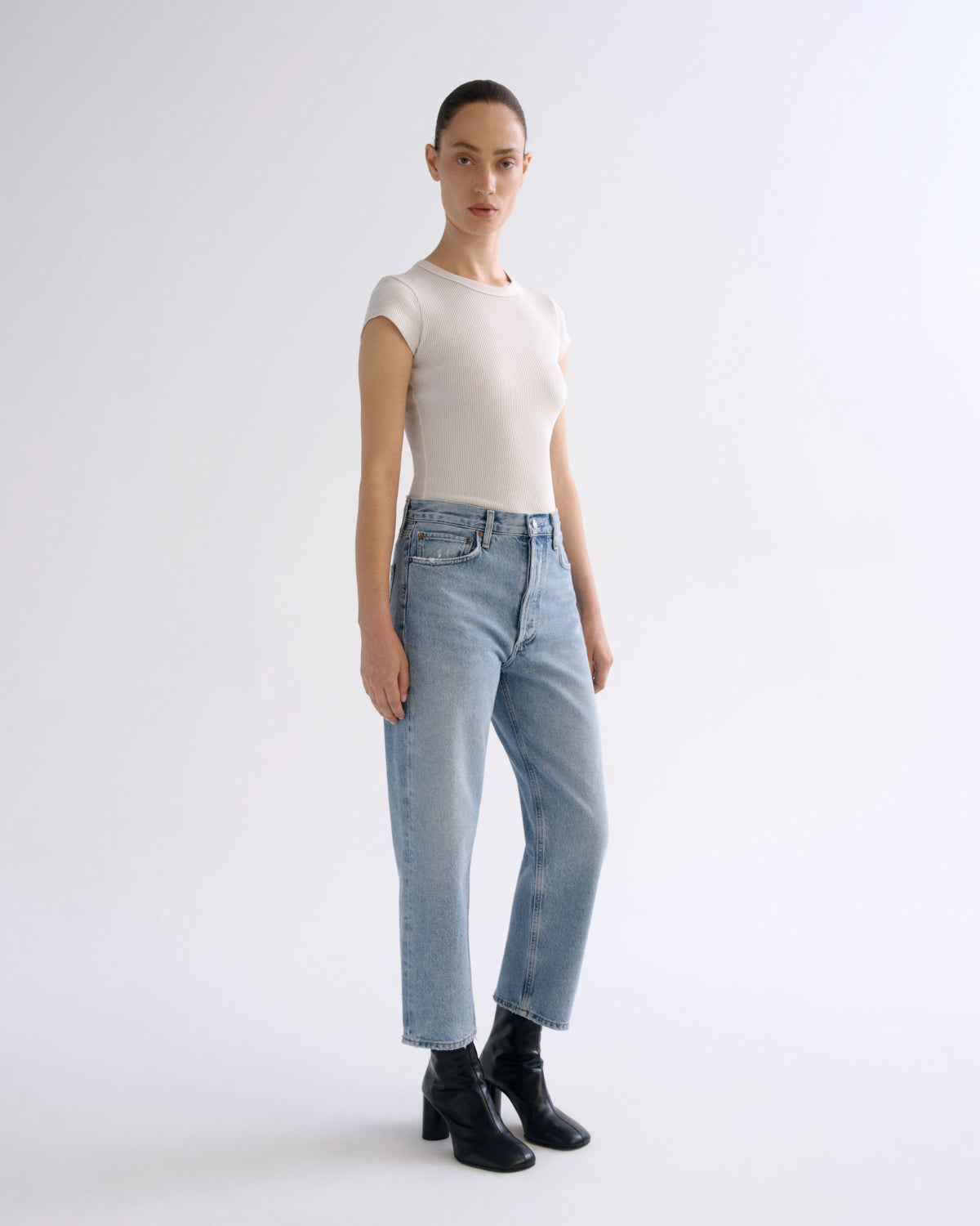 A model stands in a studio wearing a fitted cream short-sleeve top, light-wash blue jeans, and black ankle boots against a plain light gray background.