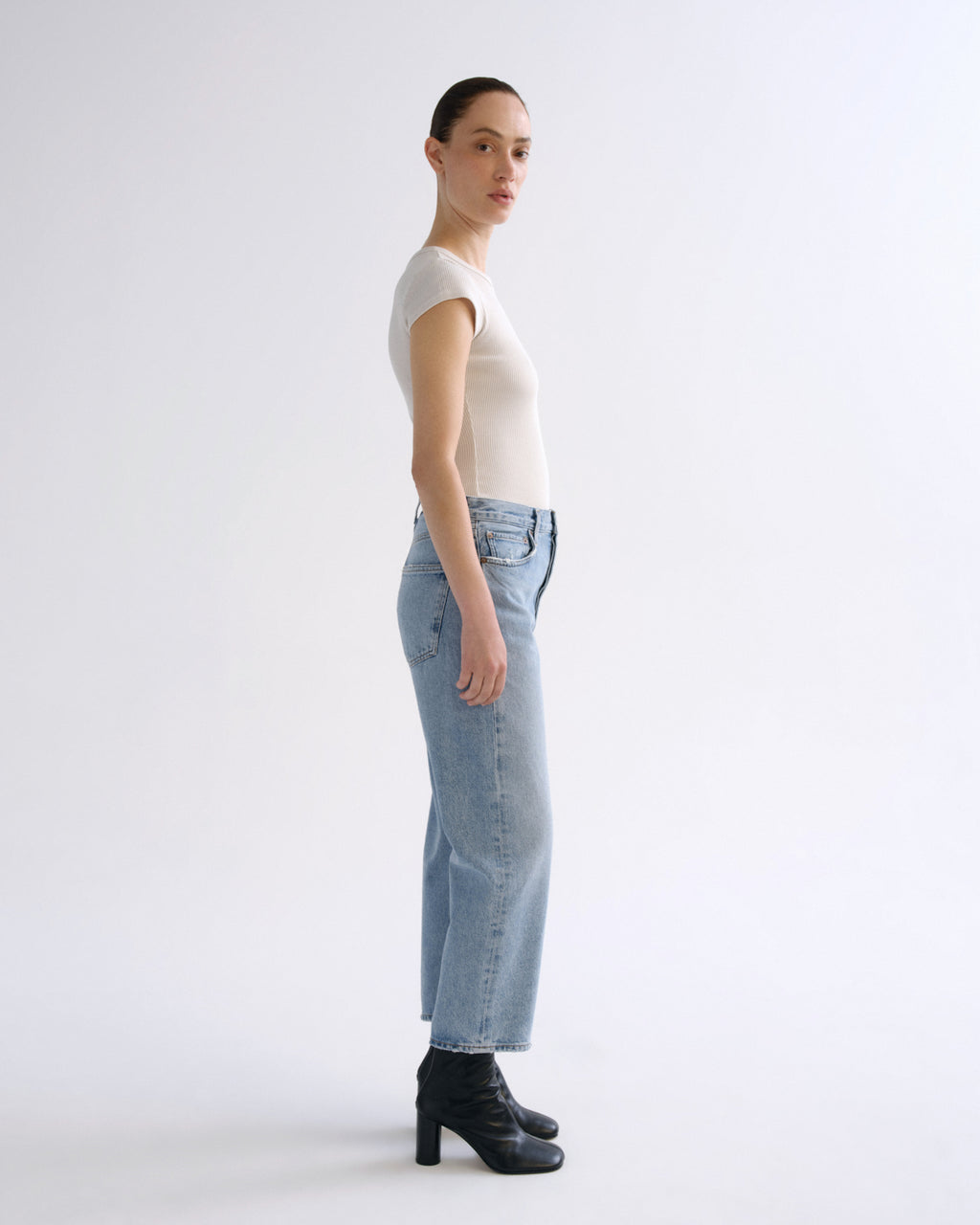 A model standing in profile wearing a fitted off-white short-sleeve top, light-wash blue straight-leg jeans, and black ankle boots against a plain light gray studio background.