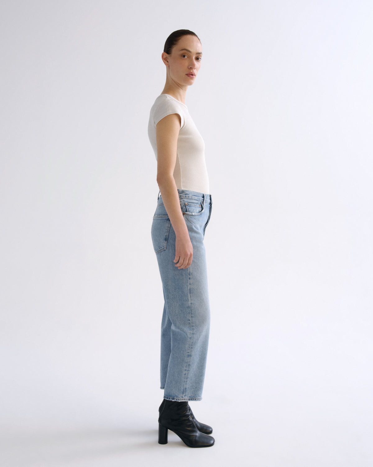 A model standing in profile wearing a fitted off-white short-sleeve top, light-wash blue straight-leg jeans, and black ankle boots against a plain light gray studio background.