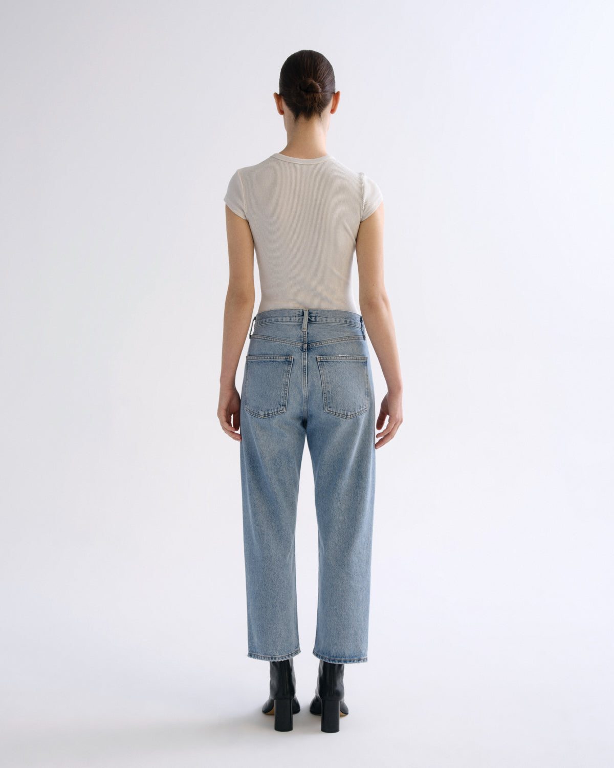 A model shown from the back wearing a light beige short-sleeve t-shirt, light-wash blue straight-leg jeans, and black ankle boots against a plain white studio background.