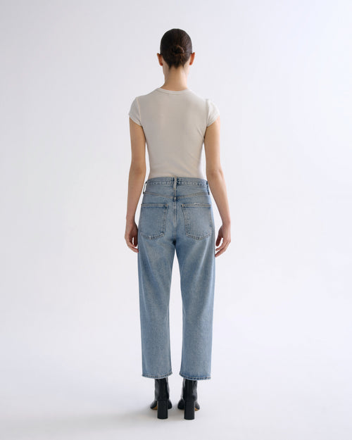 A model shown from the back wearing a light beige short-sleeve t-shirt, light-wash blue straight-leg jeans, and black ankle boots against a plain white studio background.