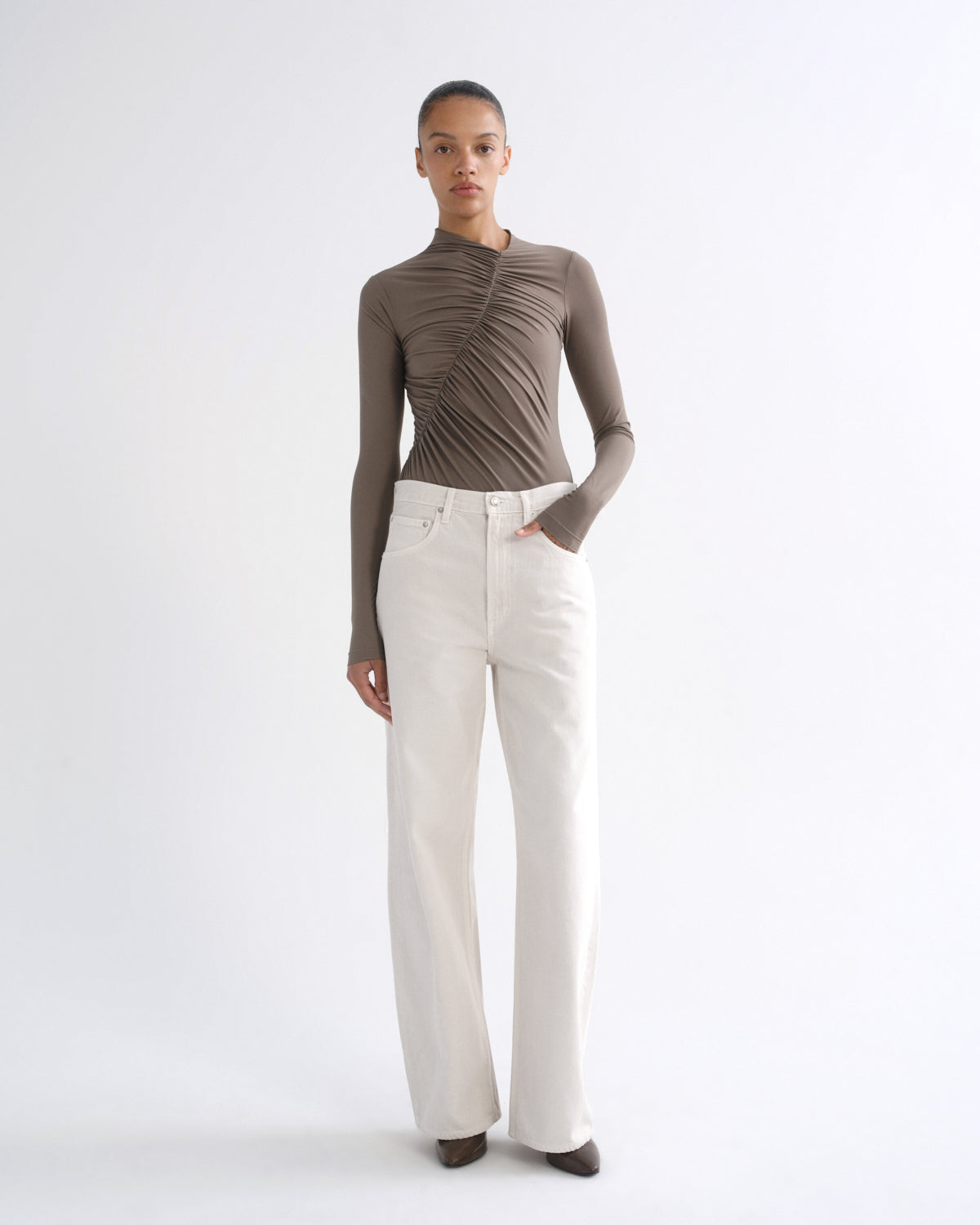 A model stands against a plain white studio background wearing a taupe-brown long-sleeve mock-neck top with dramatic diagonal pleats across the chest and white wide-leg jeans.