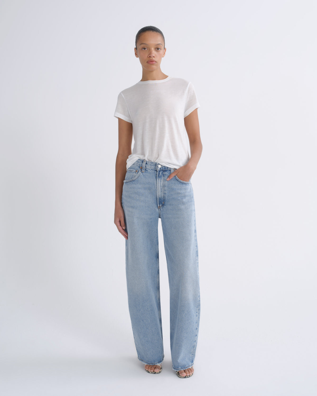 A model stands against a light gray studio background wearing a white short-sleeve t-shirt and light-wash wide-leg blue jeans.