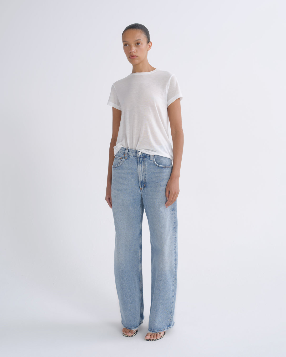 A model stands against a plain light gray studio background wearing a white short-sleeve T-shirt and light-wash wide-leg blue jeans.