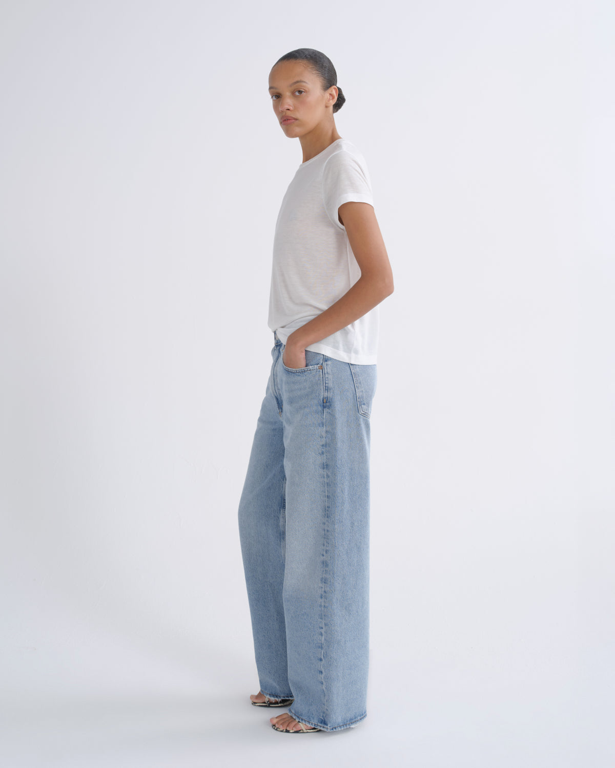 A model standing in profile wearing a white short-sleeve t-shirt and light-wash wide-leg denim jeans against a plain white studio background.