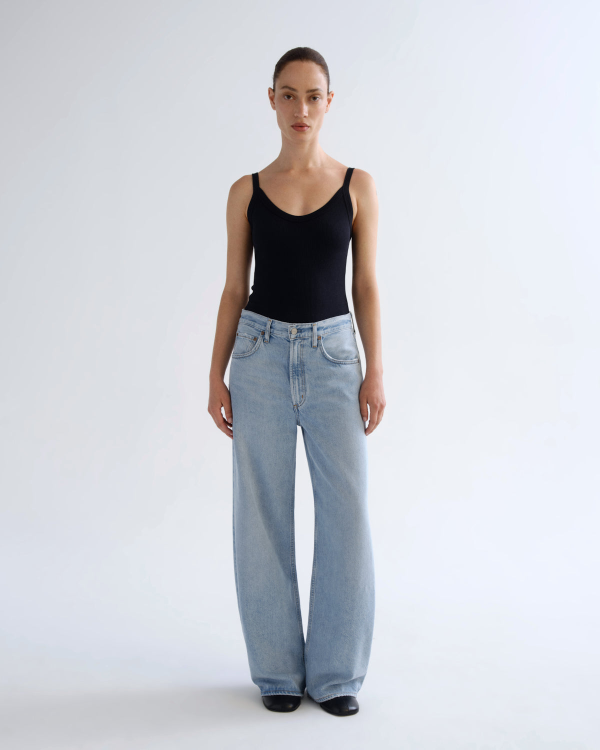 A model stands against a plain light gray studio background wearing a black sleeveless top and light-wash wide-leg jeans.