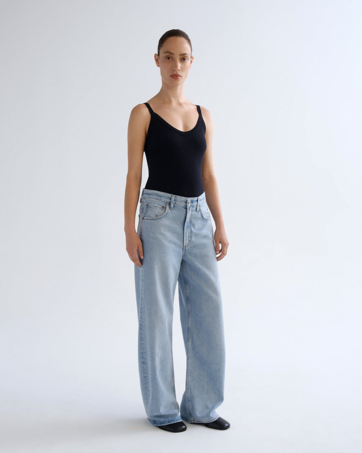 A model stands against a plain light gray studio background wearing a black sleeveless bodysuit and light-wash wide-leg jeans.
