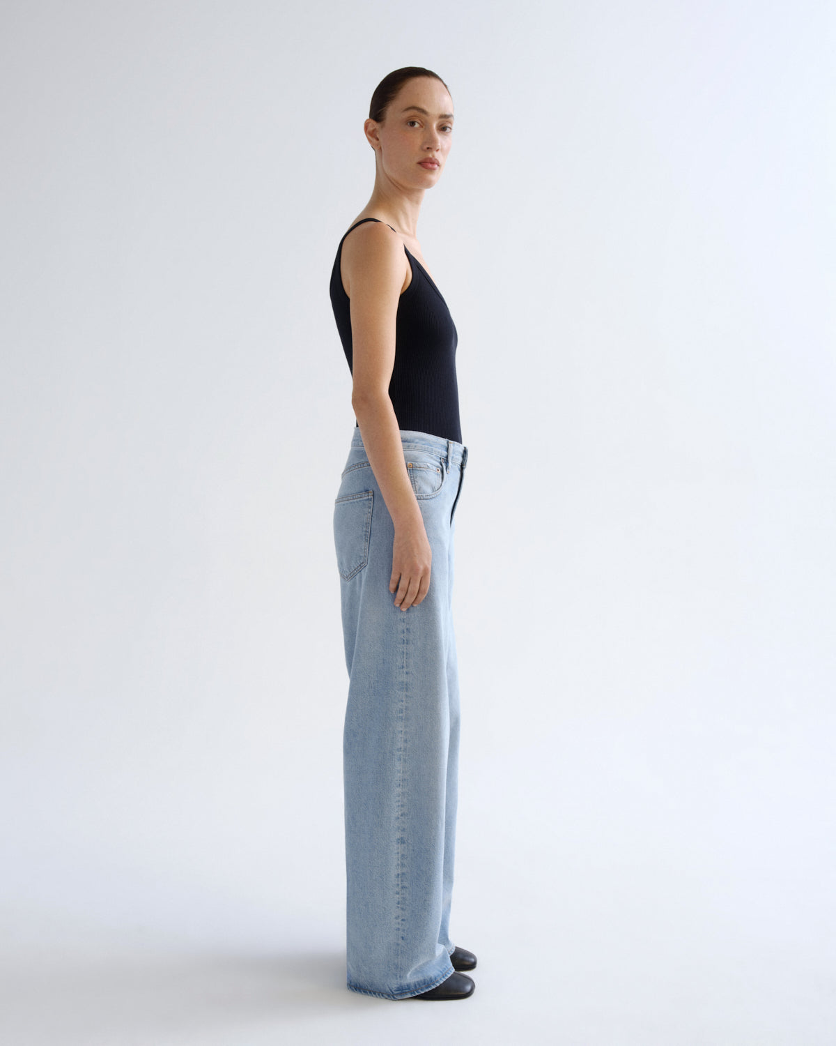 A model standing in profile wearing a black sleeveless top and light-wash wide-leg jeans against a plain white studio background.